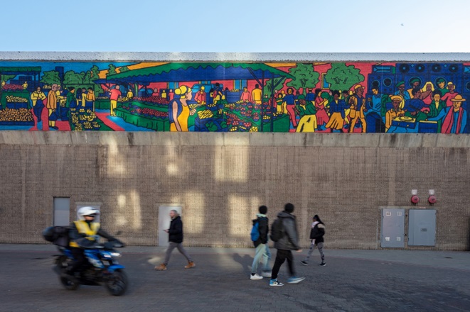 People walking past shopping centre which has a vibrant painting of the market on the buildings side.